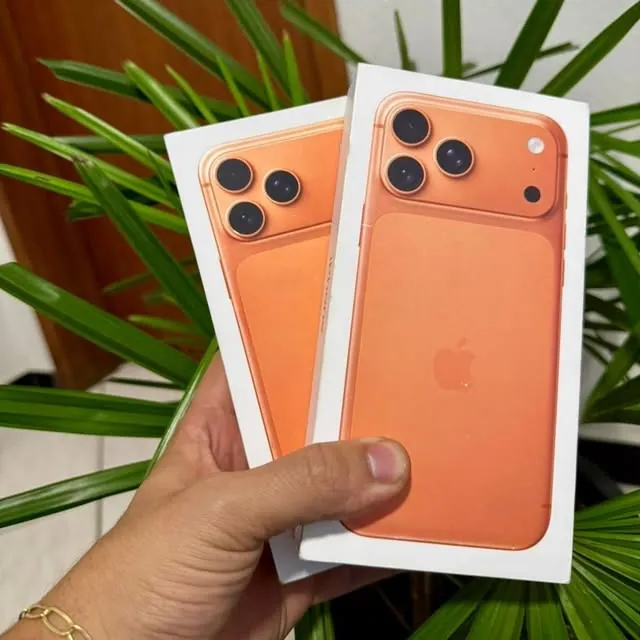 Duas caixas lacradas de iPhone 17 Pro Max laranja na mão, com folhagem ao fundo