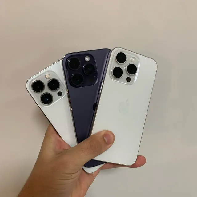 Três iPhones Pro seminovos em leque na mão, em branco e roxo-escuro
